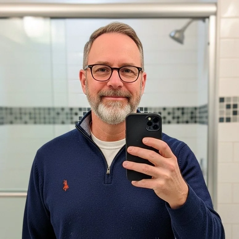 A man with glasses and a beard taking a selfie in a bathroom mirror.