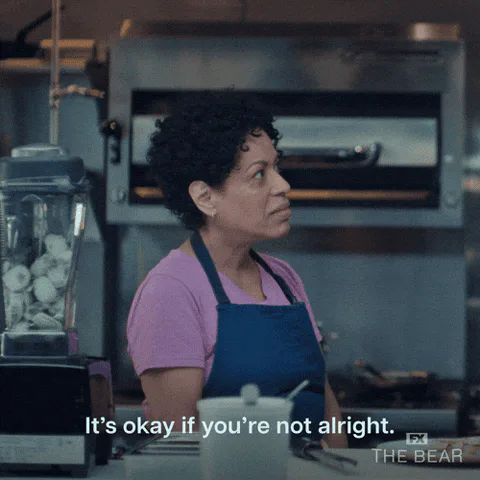 A woman in an apron in a kitchen looks aside, captioned 'It's okay if you're not alright.'