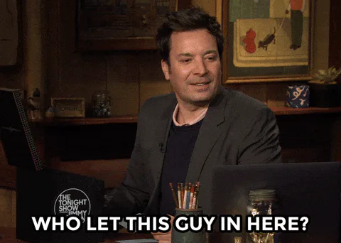 A man in a suit at an office desk smiles and says 'Who let this guy in here?'