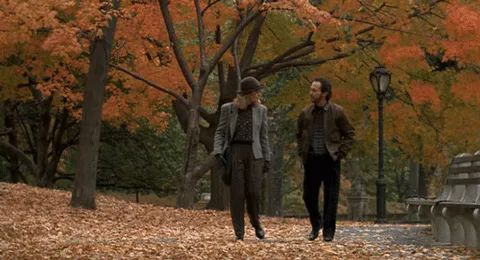 A man and a woman in coats walk together along a path covered in orange autumn leaves.