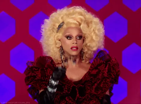 92bf9370-94bd-7e19-a003-ef248f931ced.gif A performer in a huge blonde wig, ruffled red gown and black gloves peers through jeweled opera glasses.
