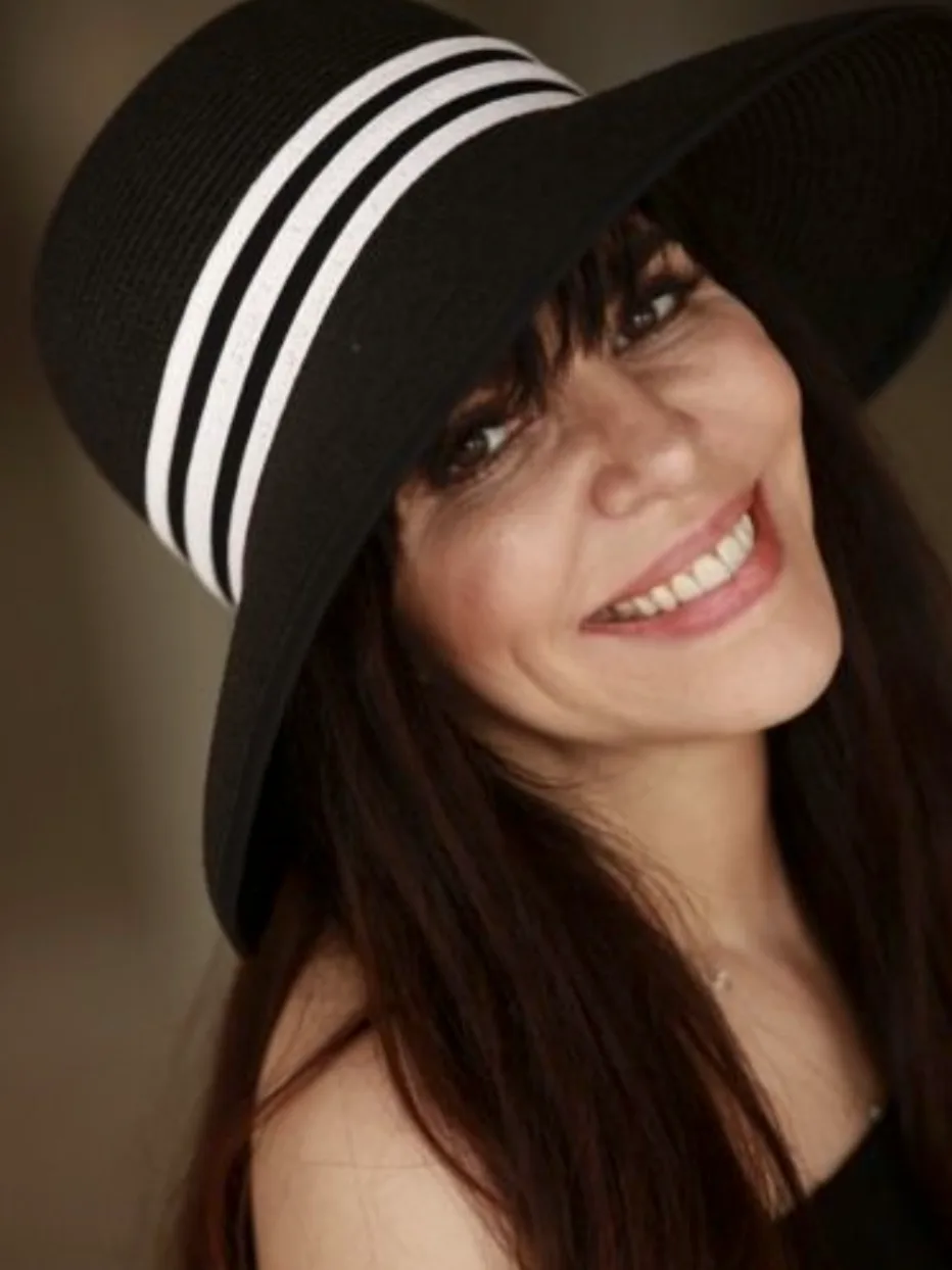 Screenshot+2023-12-14+at+12.36.34 PM.png A woman in a black sun hat with white stripes smiles, her dark hair loose over her shoulders.