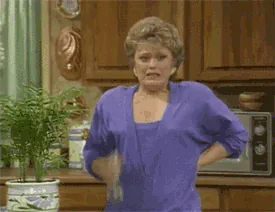 Blanche from The Golden Girls sprays water on her neck from a spray bottle in the kitchen, one hand on her hip.