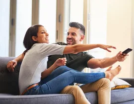 A laughing couple sits on a couch as the man reaches out, holding a remote control at arm's length.