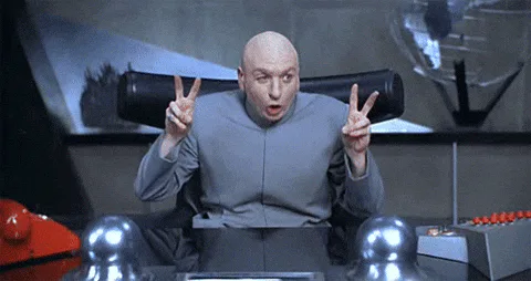 A bald villain in a gray suit makes finger gun gestures with both hands in a futuristic lair, from Austin Powers.