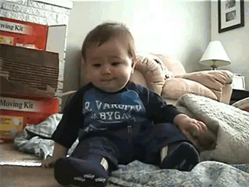 A baby sits up, tips over backward laughing, then is lifted up still laughing by an adult's arm.