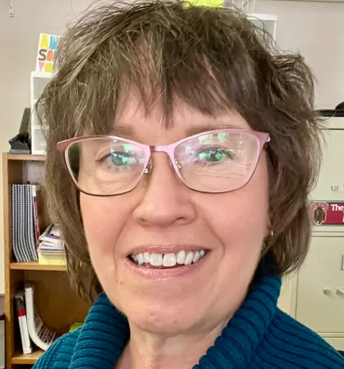 A woman with glasses smiling indoors, with books and shelves in the background.