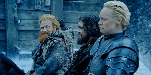A bearded man, a dark-haired man and an armored blonde woman turn to look on with unimpressed expressions.