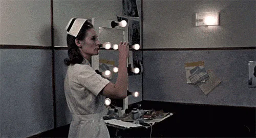 A woman in a white nurse uniform adjusts a lit vanity mirror, then a close-up shows her heavy eye makeup and a syringe.