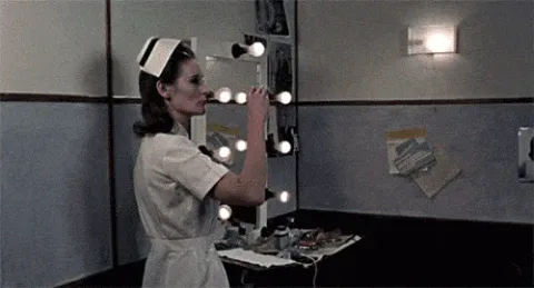 Torture+Garden+Nurse+GIF.gif A woman in a white nurse uniform adjusts a lit vanity mirror, then a close-up shows her heavy eye makeup and a syringe.