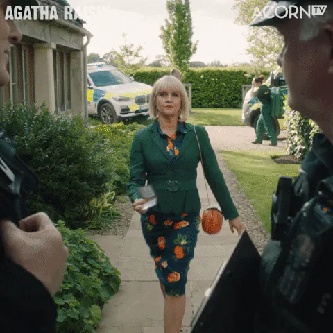 A blonde woman in a green suit holds up a phone toward two police officers outside a house.