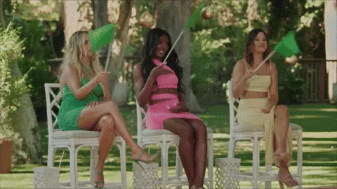 Three women in green, pink and cream outfits sit outdoors waving small green flags.