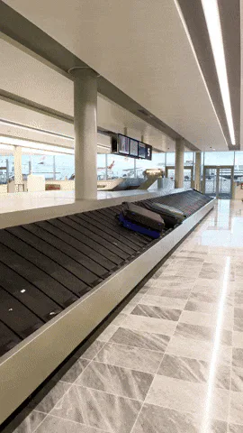 Suitcases fly off an airport baggage carousel and pile up in a chaotic heap on the floor.