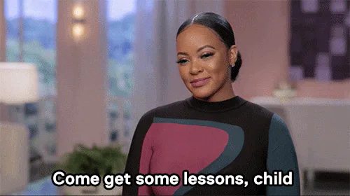 A woman with her hair pulled back waves and says 'Come get some lessons, child.'
