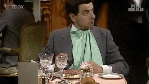 4e6bcfc7-9e60-5e23-b536-555310df1ab5.gif A man in a formal suit and green scarf sits at an elegant dinner table, pulling exaggerated faces.