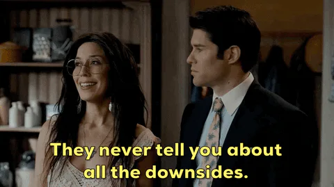 A woman in glasses talks to a man in a suit in a rustic shop and says 'They never tell you about all the downsides.'