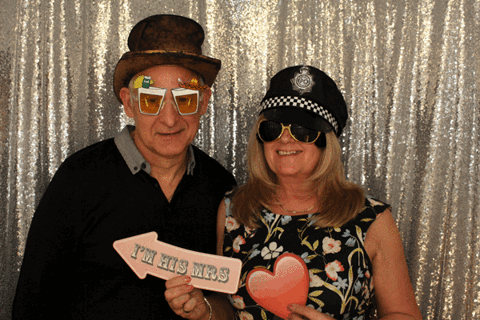 An older couple pose with a heart cutout and an 'I'm his Mrs' sign against a sparkly backdrop.