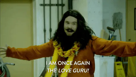 A man with a long beard and flower lei throws his arms open, declaring 'I am once again the love guru!'