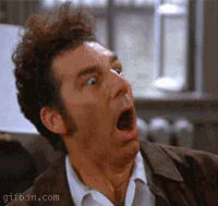A curly haired man opens his mouth wide in an exaggerated, shocked reaction.