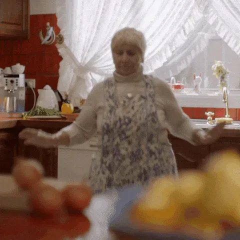 Dance+Love+GIF+by+SimAust+(1).gif An older woman in a floral apron dances in a rustic kitchen with a curtained window behind her.
