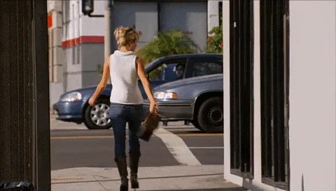 A woman in a white top and jeans walks away from the camera past a parked car on a sunny street.