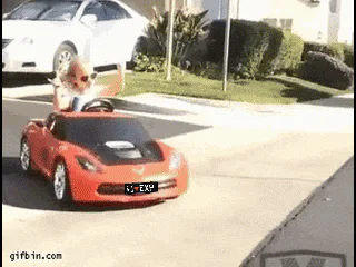 A toddler in sunglasses waves both arms while riding in a toy sports car outdoors.