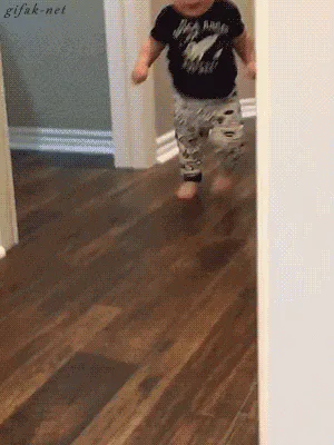 A toddler in a black space-themed shirt and patterned pants runs and stumbles down a hallway.