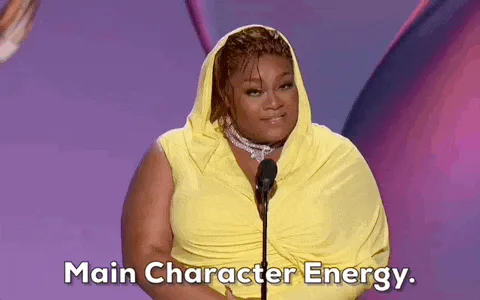 A woman in a yellow draped gown speaks at a microphone as text reads 'Main Character Energy.'