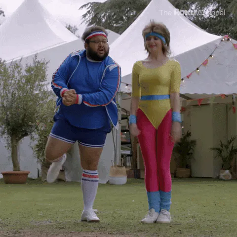 A man and woman in retro 80s workout outfits dance outdoors in a yard as text reads 'Let's go!'