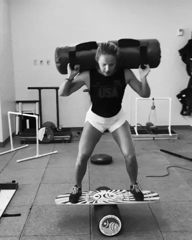 A woman in gym gear balances on a wobble board while carrying a heavy sandbag across her shoulders.