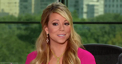 A woman with wavy blond hair sits in an office chair, smiling as she talks with a city skyline behind her.