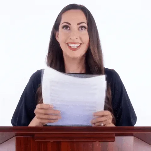 A smiling woman holds up a sheet of paper while standing at a wooden podium.