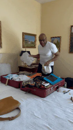 A bald man tosses a folded white item into an open suitcase on a bed in a yellow room.
