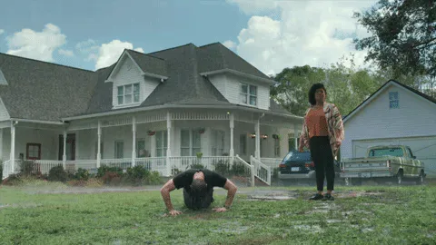 ce8b1e92-a24e-693e-e3ea-e8ed8b101d43.gif A man does push-ups on the grass while a woman stands watching in front of a farmhouse.