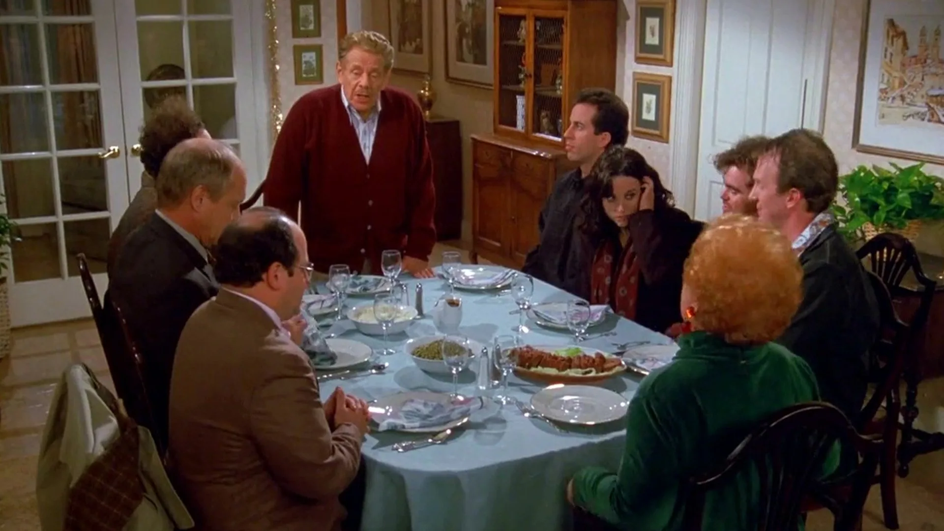 A large family gathers around a set dinner table as an older man in a red cardigan stands and speaks.