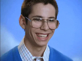 A young man in glasses and a cardigan grins widely, then smirks, in a retro sitcom clip.