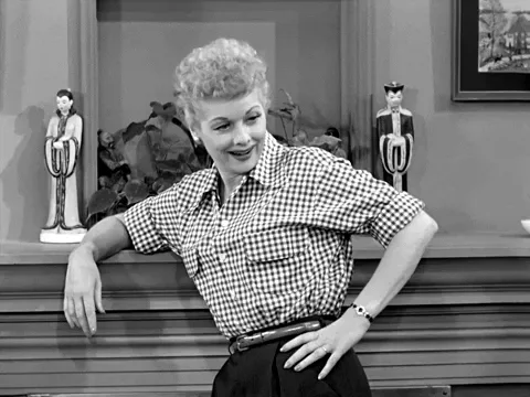 A woman in a checkered shirt leans over laughing uncontrollably, in a black-and-white sitcom clip.