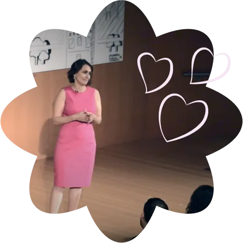 Laurie Gerber speaks on a stage in a pink dress, framed by a flower shape with heart line drawings.
