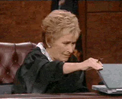 A judge in black robes points a pen at a laptop on the bench, looking unimpressed.