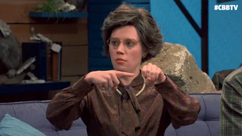 A woman with curly hair and glasses in a vintage dress makes an exaggerated pointing hand gesture.