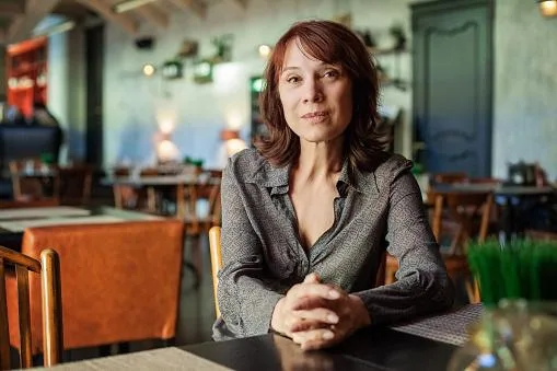 image-01-rId6.png A woman sits with her hands clasped at a table in a warmly lit restaurant.