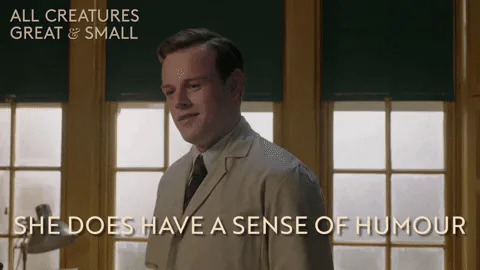 A young man in a beige suit smiles by a window and says 'She does have a sense of humour.'