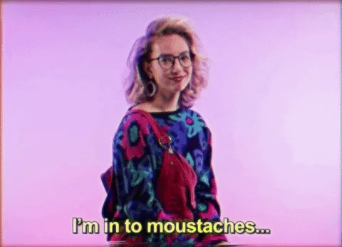 A woman with glasses and a floral top says 'I'm in to moustaches...' against a pink backdrop.
