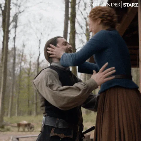 Season+6+Love+GIF+by+Outlander.gif A man and woman share a warm embrace outdoors, then hug tightly at a cabin doorway, both smiling.