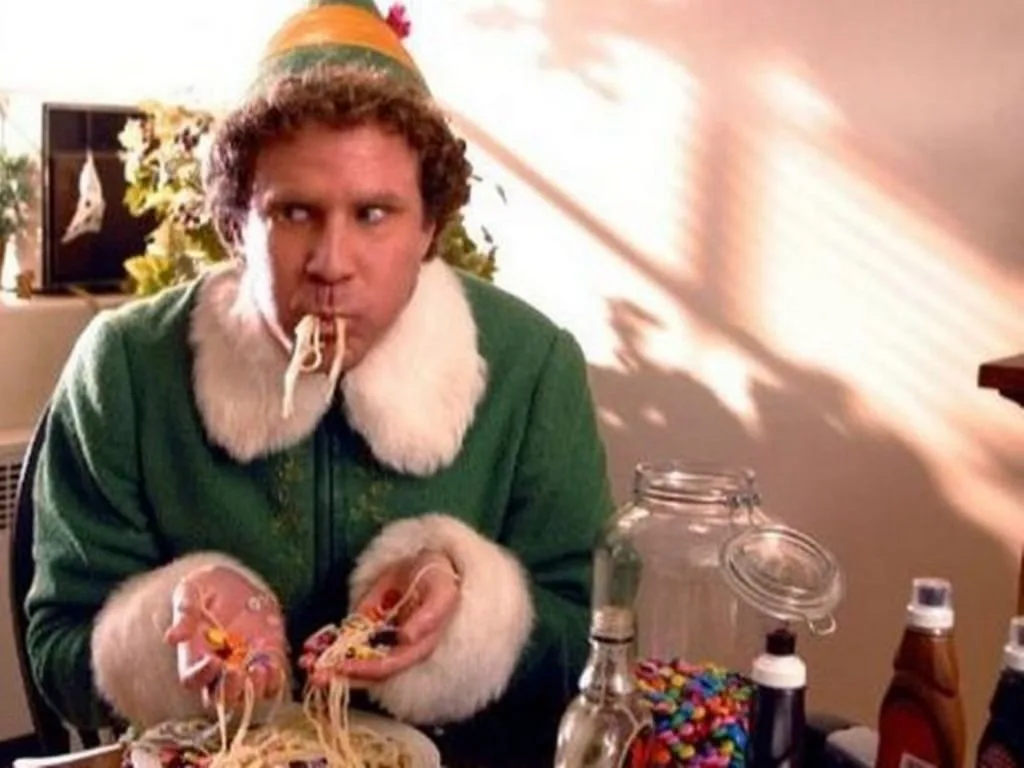 5fe56995-0a54-82e7-c61e-d6a8a3618f7b.jpeg A man in a green fur-trimmed costume eats spaghetti mixed with candy and syrup at a table.