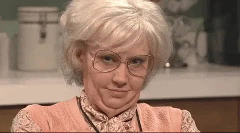 An older woman in glasses and a pink blouse winks and smiles playfully in a kitchen.