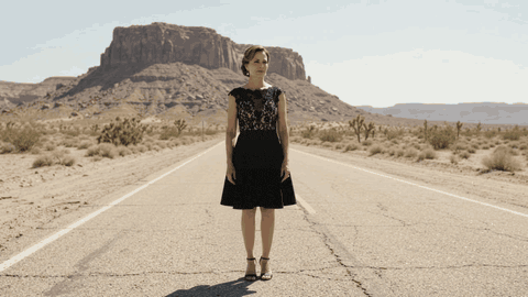 A woman in a black dress watches a tumbleweed roll across an empty desert road.