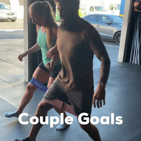 A man and woman do lateral band walks together indoors in a gym with an open garage-style door, captioned 'Couple Goals.'