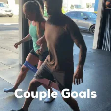 download.gif A man and woman do lateral band walks together indoors in a gym with an open garage-style door, captioned 'Couple Goals.'
