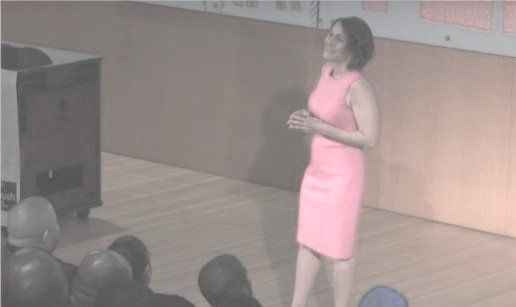 A woman in a pink dress giving a presentation on stage to an audience.